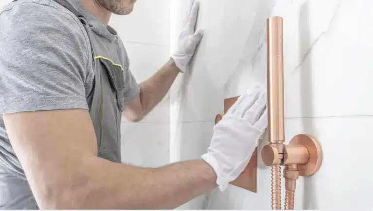 A person wearing white gloves and a grey uniform is installing a copper shower fixture on a white marbled tile wall.