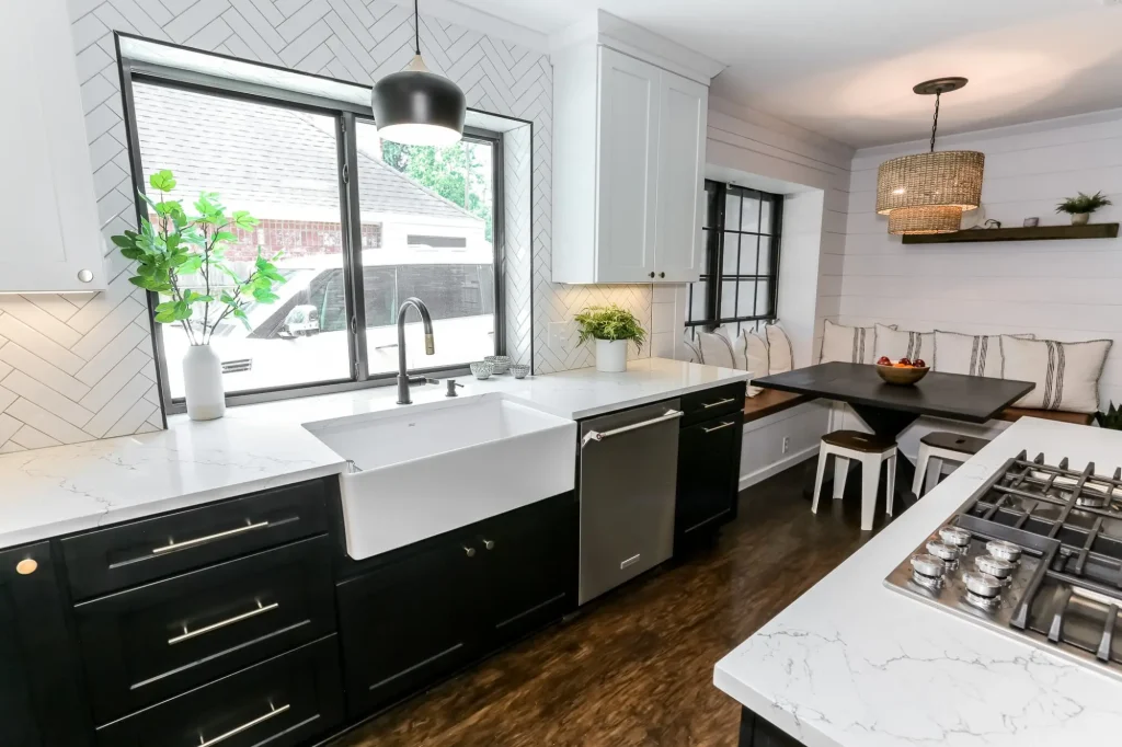 DWR Interiors kitchen renovation in Houston TX showing a white apron front farmhouse sink with brushed nickel gooseneck faucet set in black shaker cabinets with gold bar pulls, stainless steel dishwasher, white herringbone tile backsplash surrounding a large black frame window with matte black pendant light, white quartz countertop with gray veining, island gas cooktop in foreground, and built in breakfast nook banquette with woven drum pendant visible in the background