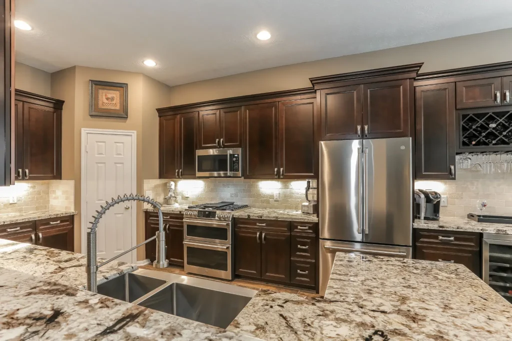 DWR Interiors kitchen renovation in Houston TX showing a granite topped island with commercial spring faucet and stainless steel undermount sink, stainless double oven gas range, over the range microwave, stainless French door refrigerator, dark cherry raised panel cabinets with crown molding, travertine backsplash with under cabinet lighting, built in wine rack with stemware holder, and pantry door