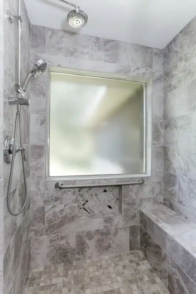 nterior of a walk in shower with ceiling mounted rain showerhead, chrome handheld shower on slide bar, brushed nickel grab bar, herringbone mosaic tile niche, built in bench, and frosted privacy window in a Houston TX bathroom remodel by DWR Interiors